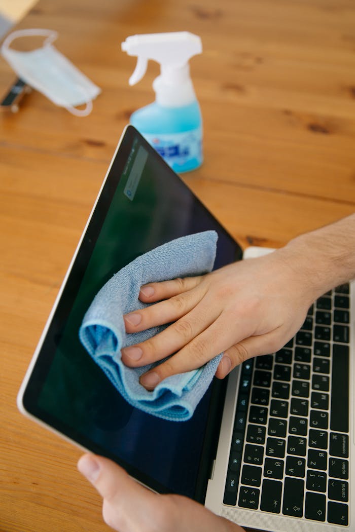 about-02 A person wipes a laptop screen using a blue cloth and spray bottle on a wooden table for hygiene.